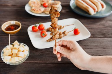 Bir el ahşap bir masanın üzerinde bar aperatifleriyle ızgara tavuk şişi tutuyor.