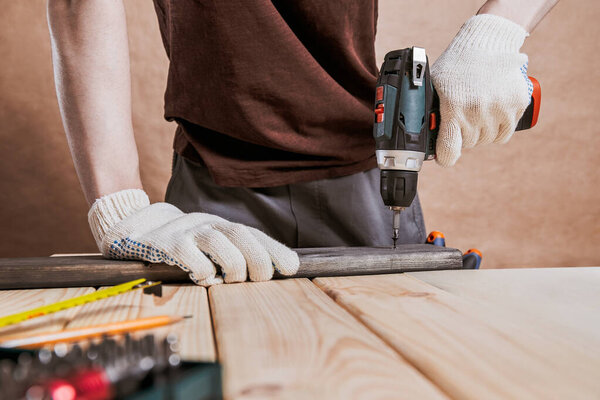 Person in gloves drills into a wooden beam with a cordless power tool on a workshop table, DIY repair, woodworking