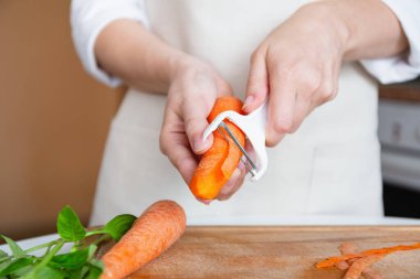 Önlüklü bir insan, elinde sebze soyacağı olan çiğ bir havucu soyar, yemek hazırlar.