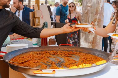 Paella festivalinde büyük bir tavadan taze paella kepçesiyle beyaz bir tabağa koyan bir şefin yakın plan fotoğrafı. 05.06.2023