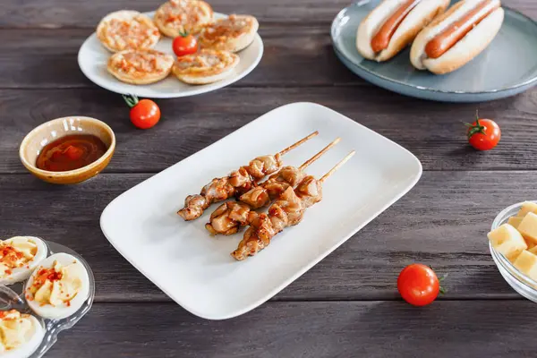 Beyaz bir tabakta ızgara yakitori tavuk şişleri, baharatlı yumurta, peynir küpleri ve atıştırmalıklarla çevrili. Bira, parti yemeği.