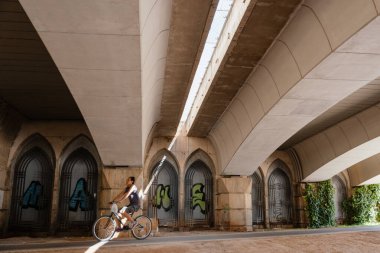 Bir adam büyük şehir tarzı köprünün altında bisiklet sürüyor, güneş ışığı, şehir bisikleti, sokak hayatı - Valencia, İspanya, 05.06.2023