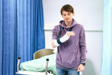 Kırık kol askıda, mavi perdeli hastane yatağının yanında duruyor.