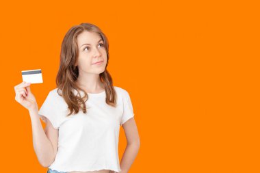 Elinde kredi kartı olan beyaz gömlekli genç bir kadın turuncu arka planda dalgın bir ifadeyle internetten alışveriş yapıyor.