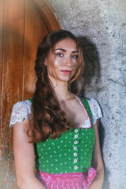 Resim, eski bir ahşap kapının ve taş bir duvarın önünde duran geleneksel bir Bavyera dirseği içindeki genç bir kadını gösteriyor. Onun uzun, kahverengi saçları omuzlarının üzerinden yumuşak dalgalar halinde düşüyor. İnce bir desenle süslenmiş dirndl 'ın yeşil korsesi.