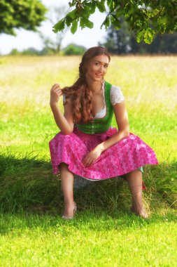 Bu resim, geleneksel bir Bavyera çayırında mutlu bir şekilde oturan genç bir kadını gösteriyor. Yeşil bir korse giyiyor ve pembe bir önlüğü var. Renkleri çayırın yemyeşil renkleriyle uyumlu. Kadın arkadaşına gülümsüyor.