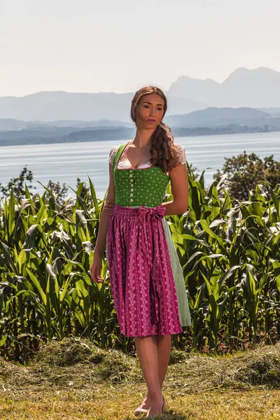 stock image The picture shows a young woman wearing a traditional Bavarian dirndl and standing in front of a picturesque natural backdrop. A tranquil lake stretches out in the background, framed by rolling hills and majestic mountains. The woman is wearing a gre