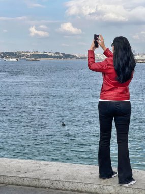 Türkiye 'nin İstanbul kentinde, cep telefonu veya cep telefonuyla şehir manzarasının fotoğrafını çeken genç, zayıf bir turistin arkası. Deniz kenarında cep telefonlu bir kadın.