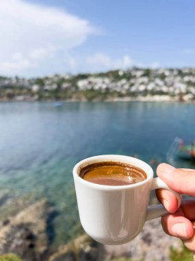 Türkiye 'de güneşli bir yaz gününde, deniz ve adanın üzerinde bir fincan Yunan ya da Türk kahvesi tutan kahve, yakın plan kadın eli. El, deniz ve gökyüzünde fotokopi uzayı ile espresso bardağını tutuyor. sıcak içecek