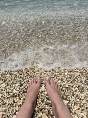 Bare feet on pebbly beach shore with gentle sea foam, relaxing summer moment, clear coastal water, shoreline tranquility, vacation and barefoot beach therapy concept