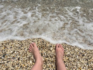 Bare feet on pebbly beach shore with gentle sea foam, relaxing summer moment, clear coastal water, shoreline tranquility, vacation and barefoot beach therapy concept