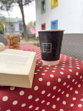 Side view paper Take away Tea or Coffee Cup on Polka Dot Tablecloth with opened Book, Biscuits over colorful Urban Courtyard. Charming outdoor coffee scene with book, black paper cup with copy space