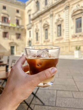 coffee, side view hand holds glass of cold coffee known as leccese in Lecce, Puglia region of Italy. traditional cold drink or beverage of Lecce in Italy. glass of cold drink over city with copy space