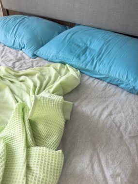 Yatak, buruşuk yeşil battaniye ve mavi yastıklı kırışık gri çarşaf, gündelik dağınık yatak odası, rahat ve gerçekçi yatak odası sahnesi yumuşak gün ışığı ve rahat atmosfer. Düzensiz yatak odası arka planı