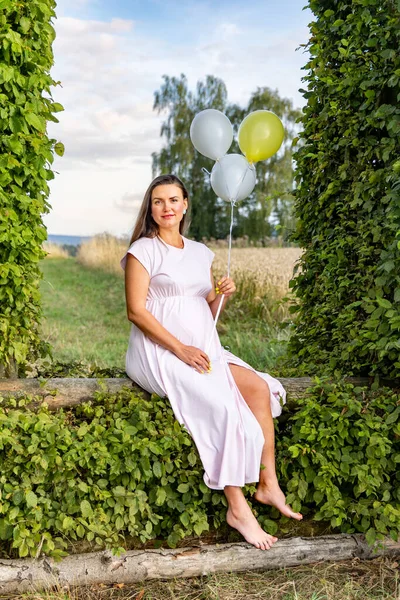 風船を持った妊婦が緑の葉でできたアーチに座っている — ストック写真