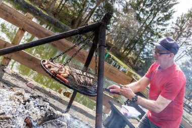 Bardaklı yaşlı bir adam dışarıda ızgarada sosis pişiriyor.