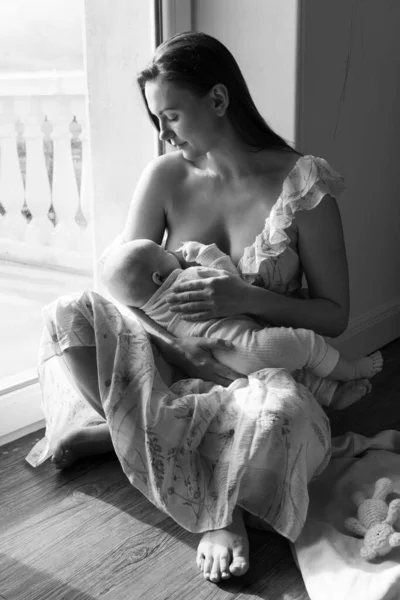 一个女人坐在窗边的地板上给她的孩子喂奶 小孩吸母乳 呵护柔情保护 母爱的概念 天然营养 单色照片 — 图库照片