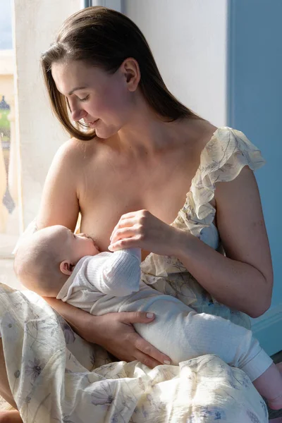 一个女人坐在窗边的地板上给她的孩子喂奶 小孩吸母乳 呵护柔情保护 母爱的概念 天然营养 — 图库照片