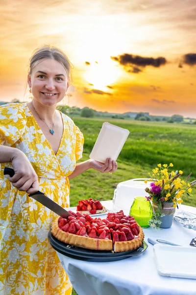 A woman cuts a cake for her birthday against the background of the sunset
