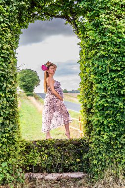 Elinde çiçekle hamile bir kadın yeşil yapraklardan yapılmış bir kemerde duruyor.