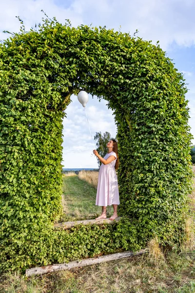 A pregnant woman with balloons is standing in an arch made of green leaves