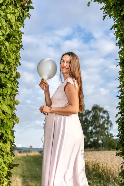 A pregnant woman with balloons is standing in an arch made of green leaves