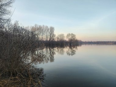Nehirde akşam manzarası ve suda yansıması var. Renkli bulutlu gökyüzü ve ağaçlar sakin nehir suyuna yansıyor. Çimenler ve ağaçlar nehrin kıyısında yetişir. Evler suya yansıyor..
