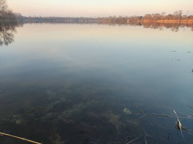 Nehirde akşam manzarası ve suda yansıması var. Renkli bulutlu gökyüzü ve ağaçlar sakin nehir suyuna yansıyor. Çimenler ve ağaçlar nehrin kıyısında yetişir. Evler suya yansıyor..