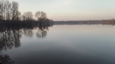 Nehirde akşam manzarası ve suda yansıması var. Renkli bulutlu gökyüzü ve ağaçlar sakin nehir suyuna yansıyor. Çimenler ve ağaçlar nehrin kıyısında yetişir. Evler suya yansıyor..