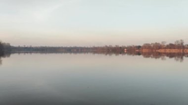 Nehirde akşam manzarası ve suda yansıması var. Renkli bulutlu gökyüzü ve ağaçlar sakin nehir suyuna yansıyor. Çimenler ve ağaçlar nehrin kıyısında yetişir. Evler suya yansıyor..