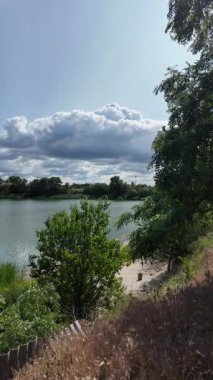 Nehrin kıyısına yürü. Göl kıyısı. Nehir kıyısında. Çimenler ve ağaçlar gölün kıyısında yetişir. Söğütlerin dalları suyun üzerinde uzanıyor. Uzak sahilde apartmanlar var..