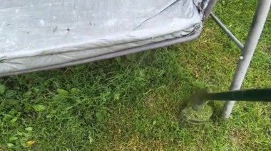 Bahçıvan uzun yeşil çimenleri biçer, erkeksi elleri tutar, çim biçme makinesi, farklı yönlerde uçan yabani otlar, yakın plan