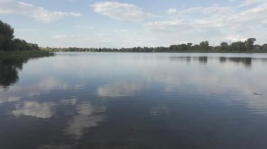 Su kıyısındaki doğal manzara. Göl kıyısı. Sudaki bulutların yansıması. Nehirle akşam manzarası. Denize doğru yürü. Su yüzeyi. Ukrayna. Düz renk profilli 4k video. D-Günlük M.