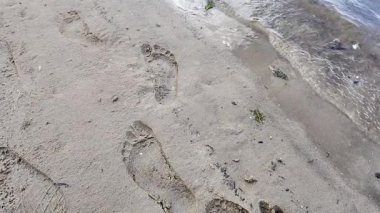 Kumda ayak izleri. Kumda su ve parmak izleri var. Kadın bacakları ya da erkek bebek, çocuk, çocuk, çıplak ayak kumsalda. Kız denize ya da okyanusa karşı dinleniyor. Su dalgaları merdivenleri yıkıyor ya da kalp şeklinde..