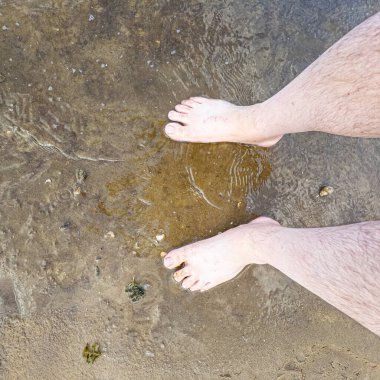 Çıplak erkek bacakları kıyıya yakın ıslak kumsalda, yumuşak dalgalar ve desenli kum tanecikleriyle. Ilık doğal ışık, sakinliği, sabit havayı arttırır, 4K