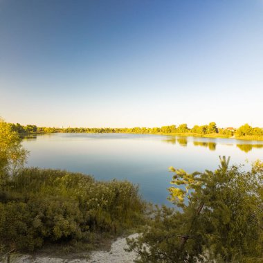 Altın güneş ışığı, bereketli sonbahar ağaçları tarafından çerçevelenmiş sakin bir nehri yıkıyor. Sakin suda yansımalar parıldıyor, bu sakin orman manzarasında huzur ve sükuneti davet ediyor..