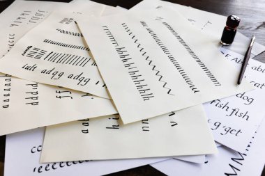 Calligraphy practice pages scattered on a table with dip pen and small bottle of ink