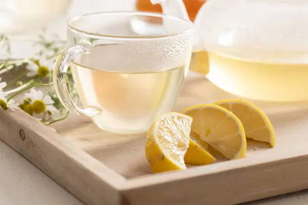 热甘菊茶在玻璃杯茶杯里 坐在一个木制盘子里 上面放着甘菊花和柠檬 免版税图库照片
