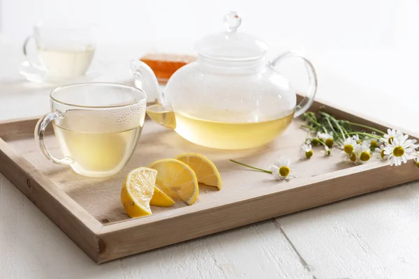 热甘菊茶在玻璃杯茶壶和茶杯里 坐在一个木制盘子里 上面放着甘菊花和柠檬 图库照片