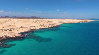 Sersemletici hava görüntüsü canlı bir okyanusu, kayalık sahili ve açık mavi gökyüzünün altındaki kumlu sahili gösteriyor..