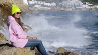 The girl sits against the background of breaking waves on the rocks. High quality 4k footage