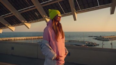 A girl walks under the roof of a building with solar panels. Pink jacket, bright hat.