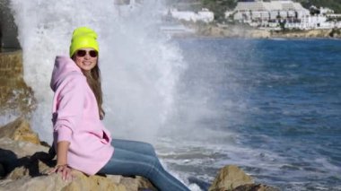 The girl sits against the background of breaking waves on the rocks. High quality 4k footage