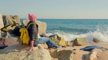 Kız kayalıklarda, deniz kıyısında, parlak güneşin altında oturuyor. Yüksek kalite 4k görüntü