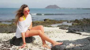 Beyazlı bir kız volkanik bir sahilde oturuyor, ayaklarının altında küçük beyaz taşlar. Yüksek kalite 4k görüntü