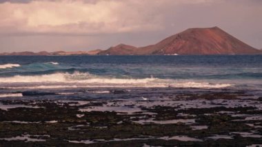 Dalgalar taştan kıyıya çarpıyor. Uzakta volkanik bir adanın siluetini görebilirsiniz. Yüksek kalite 4k görüntü