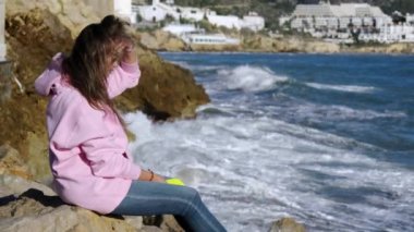 The girl sits against the background of breaking waves on the rocks. High quality 4k footage