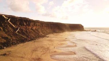 Gün batımında, resim gibi kumlu bir plajın üzerinde süzül, kamera yüksek kıyıların büyüleyici görüntüsünü yakalar, altın renklerin sıcaklığıyla kıyı manzarasının huzurlu güzelliğini harmanlar.