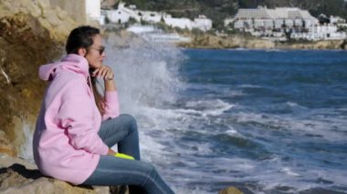 The girl sits against the background of breaking waves on the rocks. High quality 4k footage