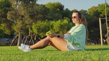 Gün batımında çimlerin üzerinde oturan bir kız, akıllı telefon kullanıyor. Yüksek kalite 4k görüntü